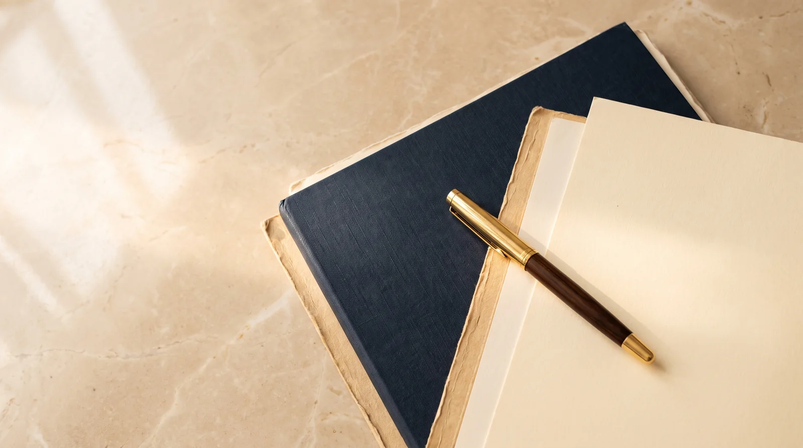 A navy document folder and cream papers with a gold pen on warm marble in soft morning light, seen from above.
